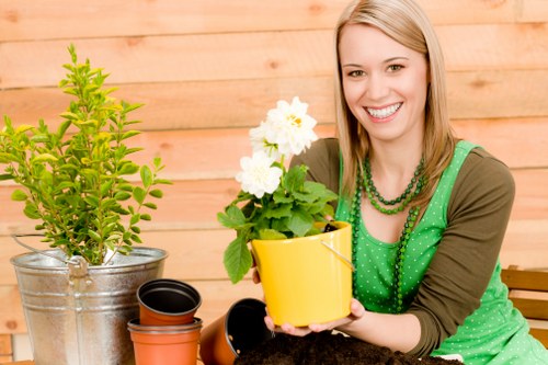 The image shows a cheerful woman with blonde hair smiling while holding a yellow flower pot containing a flowering plant with large white blossoms and green foliage, set against a background of a wooden garden fence. In front of her on the garden table are various gardening tools and containers, including a small black pot, an orange terracotta pot, and a silver metal bucket filled with fresh soil. The garden area features a well-maintained lawn with dense, lush grass, creating a vibrant green foreground. Behind her, there are contained garden elements such as potted plants and possibly a shrub or small tree in the metal bucket, indicating active gardening or potting work. The lighting suggests a bright, clear day, emphasizing the natural textures of the grass, soil, and potted plants. This scene, captured in an outdoor residential garden near Gipsy Hill, reflects a gardening service that maintains and enhances outdoor spaces with attention to plant health and garden aesthetics, aligning with local gardening practices and landscaping services offered by Gardening Services Gipsy Hill.